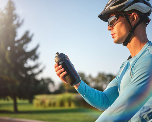 Hydration matters. Professional male cyclist holding water bottle, standing with his bike in park on a sunny day. Sport and recreation concept. Side view